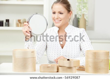 Beautiful young woman sitting at table, holding makeup mirror, smiling.