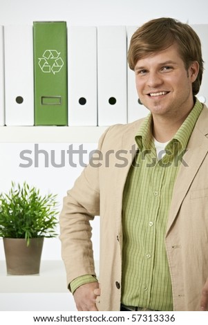Portrait of young environmentalist man posing in front of shelf in office, recycling logo on green folder.?