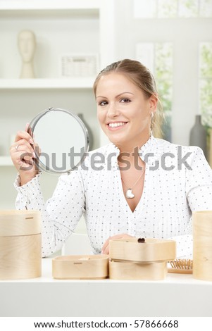 Beautiful young woman sitting at table, holding makeup mirror, smiling.