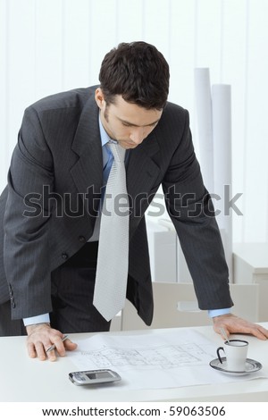 Architect wearing grey suit leaning on office desk with floor plan on it. Looking down.
