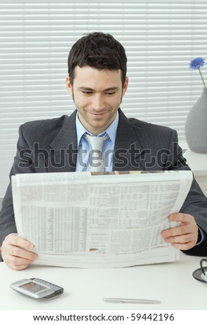 Young businessman sitting at desk and reading business news, smiling.