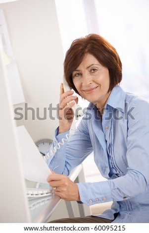 Portrait of senior office worker sitting at desk, using landline phone, holding paper.?