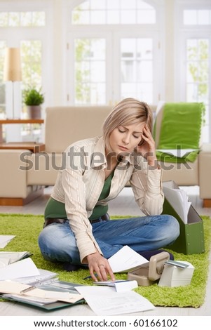 Troubled woman sitting on floor with crossed legs, doing calculation in living room.?