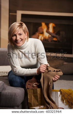 Happy young woman siiting on sofa at home taking out clothes of shopping bags. smiling.