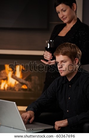 Young man sitting in front of fireplace at home working on laptop computer, woman standing behind.