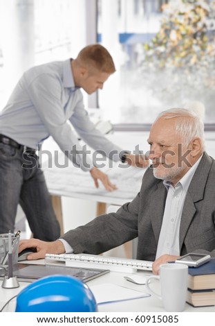 Senior businessman working on computer sitting at table, young architect standing at drawing board in background.?