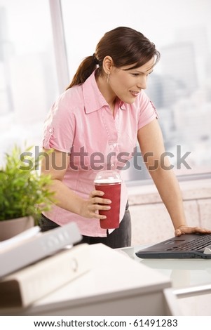 Businesswoman standing at desk holding coffee to go cup, looking at laptop.?