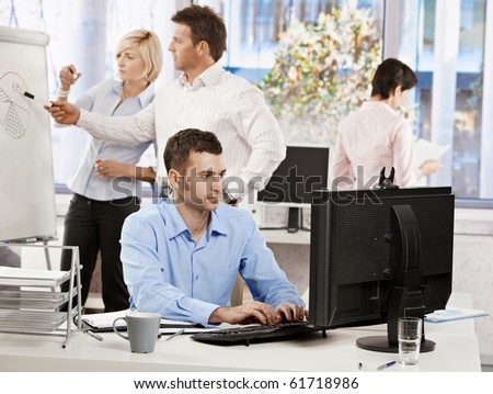 Casual businessman sitting at office desk, working on computer. Businesspeople working in background.?