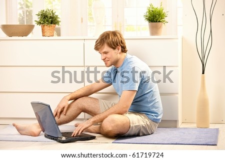 Young man browsing internet on laptop computer, typing on keyboard sitting on living room floor.?