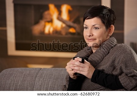 Woman sitting on sofa at home on a cold winter day, drinking hot tea, looking away.