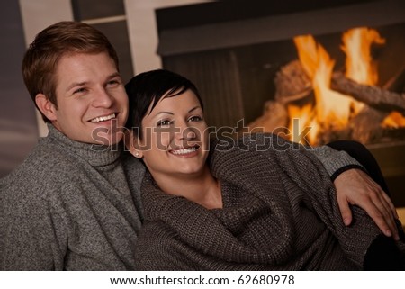 Young couple hugging in front of fireplace at home, looking away, smiling.