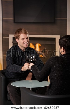 Young romantic couple dating, sitting in front of fireplace at home, drinking red wine.