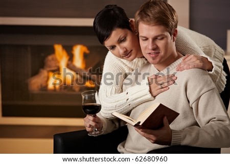 Young couple hugging in front of fireplace at home, reading book.