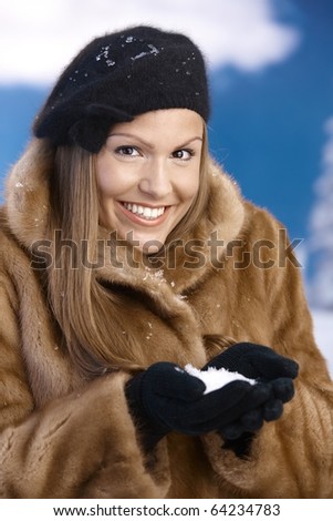 Elegant young woman dressed up warm in fur coat, cap and gloves, holding snow in hands, smiling.?