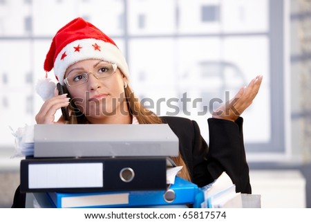 Young female in santa hat sitting in office behind folders, using mobile, having trouble.?