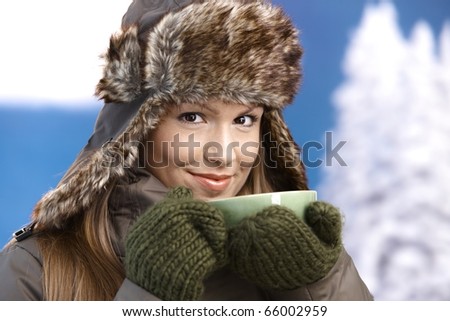 Young attractive female dressed up warm in coat, fur-hat and gloves, drinking hot tea, smiling.?