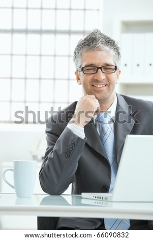 Businessman with gray heair thinking over laptop computer, sitting at desk, smiling.?