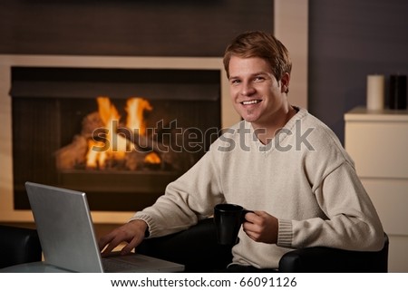 Happy young man sitting in front of fireplace at home on a cold winter day, working on laptop computer.?