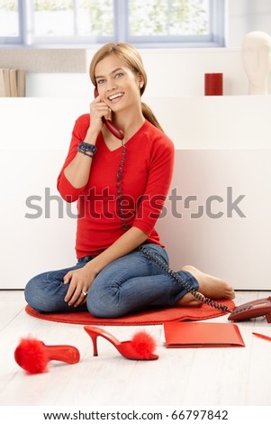 Pretty girl in red pullover talking on phone, sitting on floor at home, smiling.?