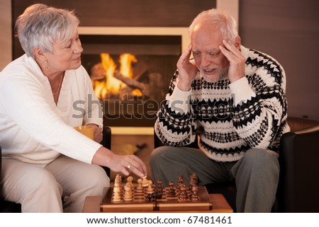 Senior couple playing chess at home, smiling woman winning game, man thinking looking troubled