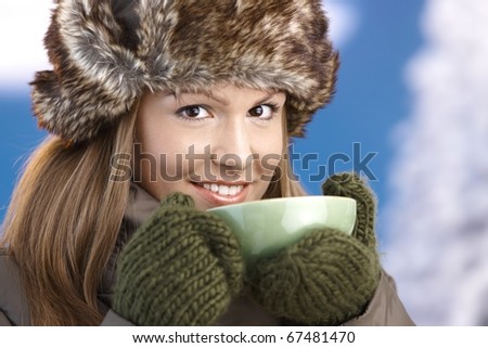 Pretty young girl dressed up warm in coat, fur-hat and gloves, drinking hot tea, smiling.