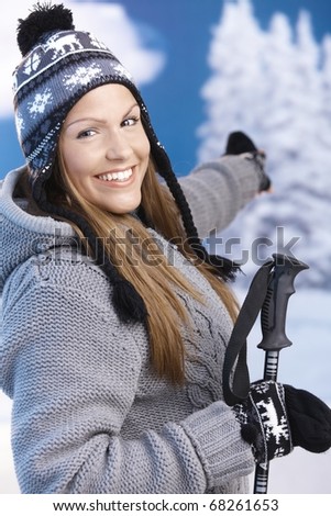 Attractive young female dressed up warm for skiing wearing cap and gloves smiling and pointing to winter landscape .?