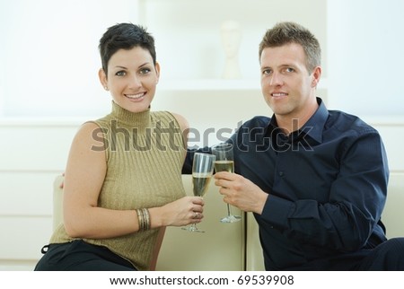 Love couple clinking champagne glasses at home on sofa. Smiling and looking at camera.