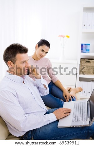 Young couple using laptop computer at home, sitting on couch. Man talkin on mobile phone.