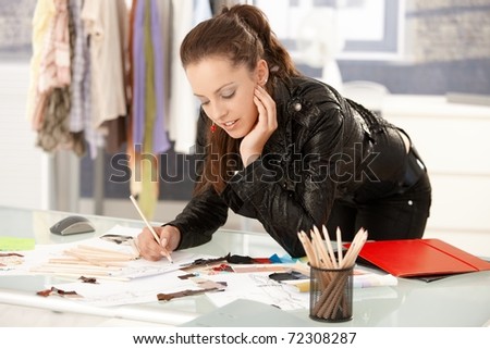 Attractive fashion designer working in office, leaning on desk, drawing.?