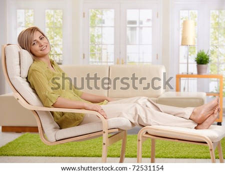 Woman relaxing at home, sitting in armchair with crossed feet up on footboard, smiling at camera.?