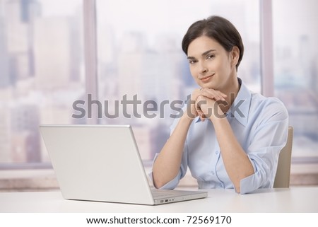 Portrait of female professional sitting at skyscraper office table with laptop computer, smiling at camera.?