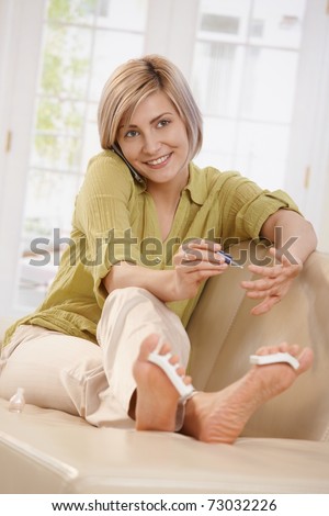 Smiling woman using nail polish on hands sitting with feet up on sofa talking on phone  in living room.?