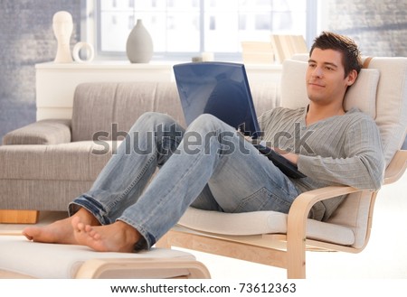 Goodlooking young man relaxing at home in armchair, sitting in living room with laptop computer, smiling.?