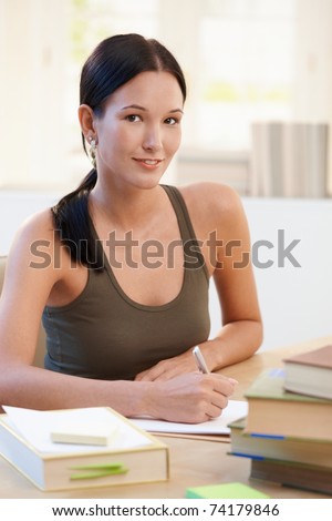 University girl studying at home, writing notes at desk, smiling at camera.?