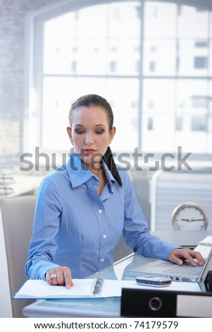 Businesswoman busy working at office desk reviewing documents and using laptop computer.?