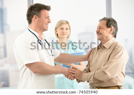 Smiling medical doctor shaking hands with happy senior patient, nurse in background.?