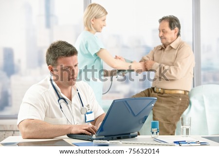 Medical checkup in doctor's office, doctor working with laptop, nurse in background measuring patient blood pressure.?