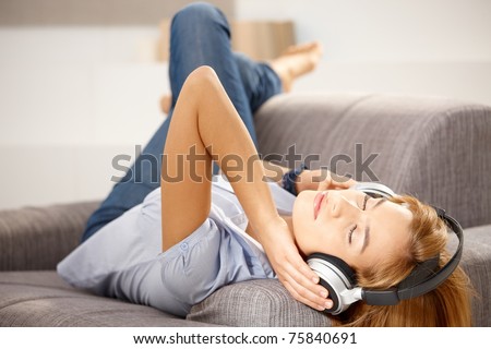 Young attractive girl enjoying music through headphones, laying on sofa.?