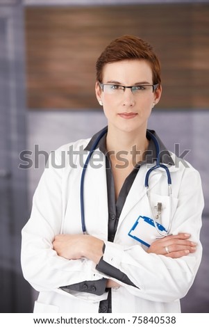 Young confident medical doctor standing in office in uniform with arms folded, smiling at camera?