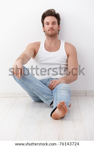 Handsome young man sitting on floor, leaning to wall, wearing undershirt and jeans.?