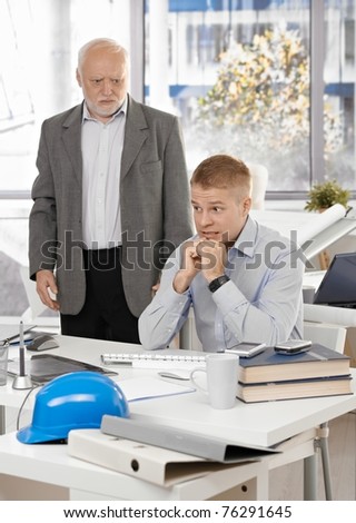 Scared young office worker man sitting at desk, angry senior executive standing.?