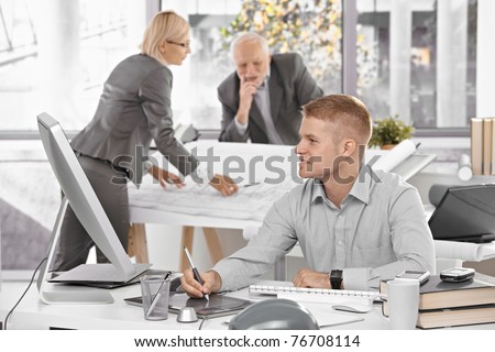 Designer team at work in office, young architect sitting at desk with drawing pad, older colleagues working on architectural plan.?