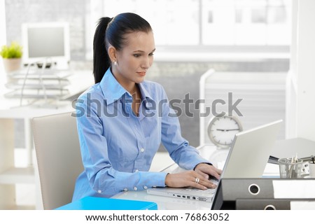 Office girl working on laptop computer at desk, looking at monitor, smiling.?