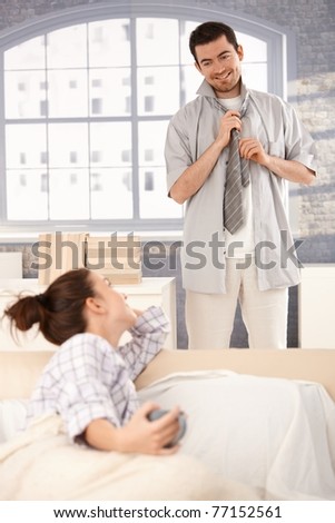 Young man dressing up in the morning, woman looking at him from bed, smiling at each other.?