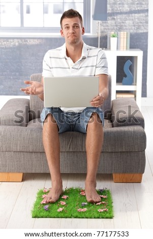 Disappointed young man sitting in armchair at home with mouth curving down, using laptop, resting legs on artificial grass.?