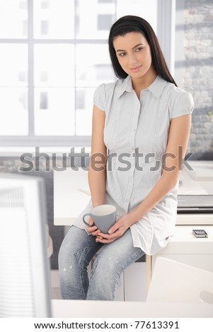 Office worker girl on coffee break, sitting on table, holding mug, smiling.?