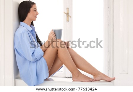Daydreaming woman wearing only shirt sitting at window sill, drinking morning coffee, smiling.?