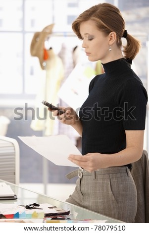 Young attractive female fashion designer standing at office desk, using mobile phone.?