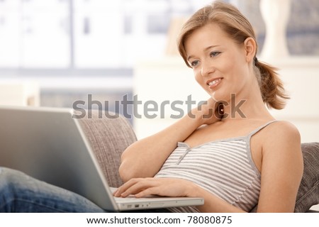 Happy young woman lying on sofa at home, browsing internet on laptop. Looking at screen, smiling.?
