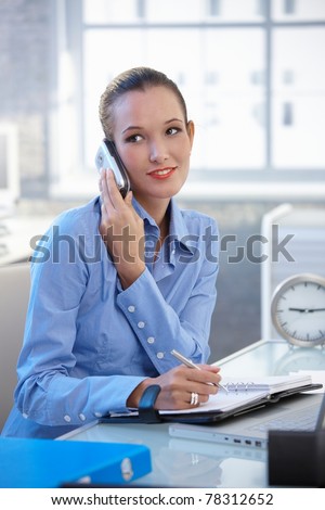 Smiling businesswoman talking on cellphone and writing into notebook at office desk.?